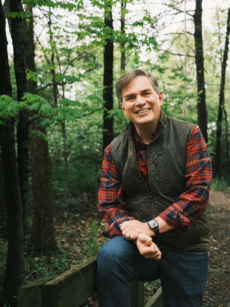 Chip Polston wearing a plaid shirt with a quilted vest over top and smiling. Diffuse trees in the background.