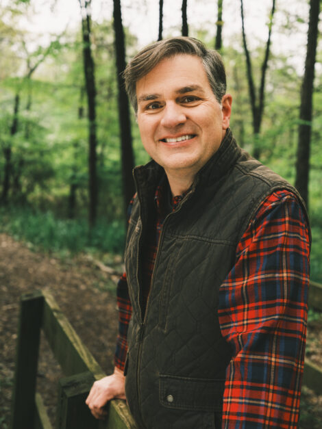 Chip Polston smiling and leaning on a wooden fence in a wooded area along a path