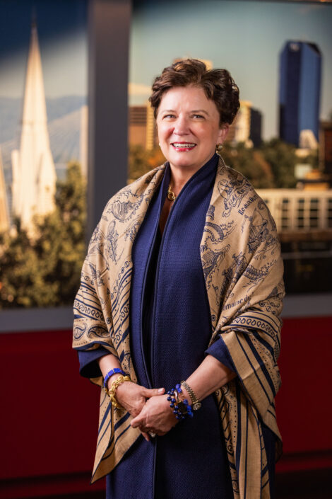 A photographic portrait of Elizabeth McCoy smiling in a blue coat with a tan and blue shawl over top.